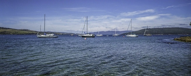 Shallow sea in the foreground with yachts moored in the water and rolling hills in the background and to the side.