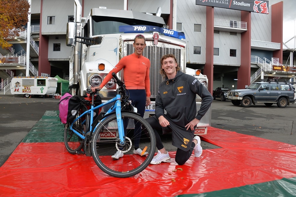 Cyclist Richie Porte stands in front of a blue and white truck next to the branded Bicycle Network ebike and kneeling next to him is Hawthorn Football Club player Josh Weddle.