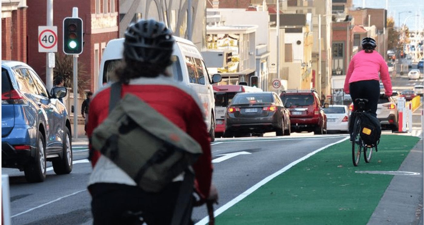 Two people ride along a green bicycle lane, the front rider in a bright pink jacket and the back slightly blurred rider wearing a red jackets with a bag slung over the shoulder.