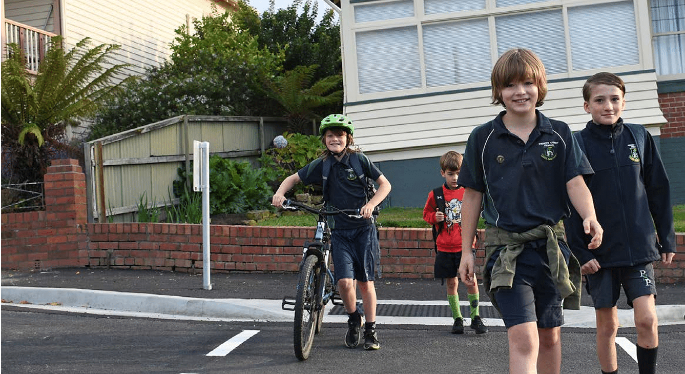 Primary school aged children cross a road, with one child wheeling a bicycle.