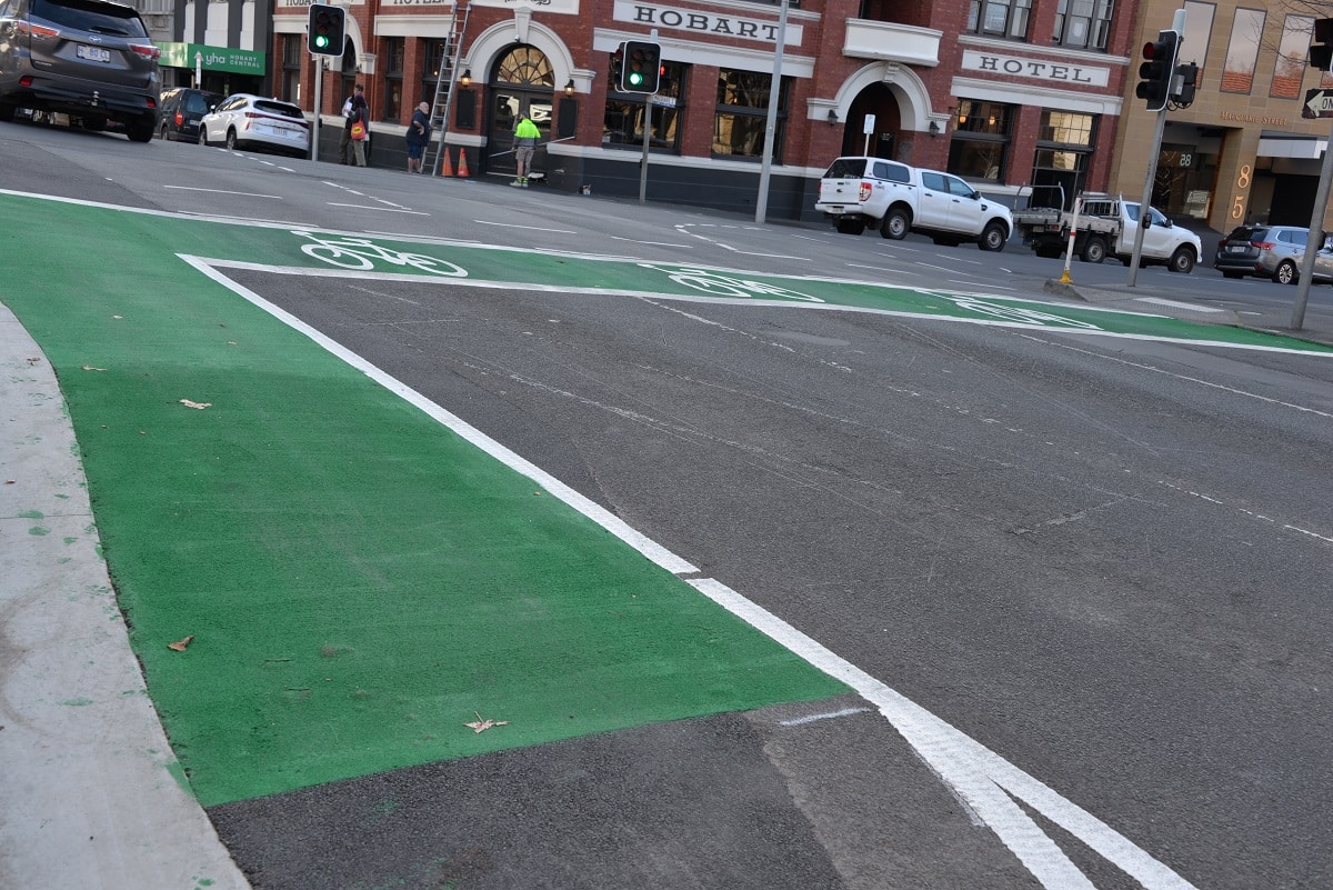 A green painted lane on the left of a road and three bicycle storage boxes with white bicycle symbols at the intersection.