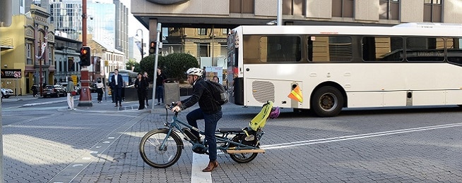 A man dressed in smart casual clothes on a cargo bike with child seat at the Collins and Elizabeth streets intersection with bus and people walking in the background.