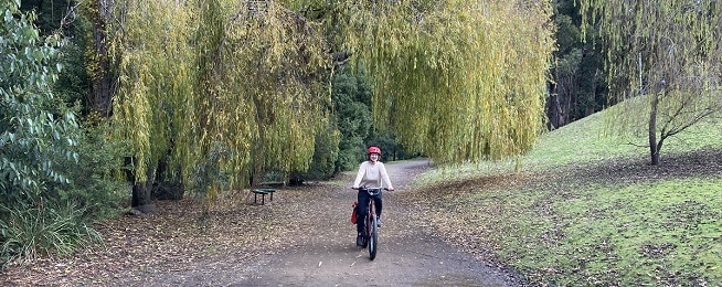 Woman in beige top and red helmet rides on a wide gravel path framed by willow trees.