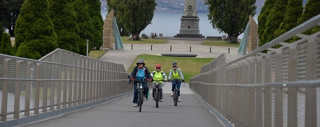 Three people on bikes ride up a steep slope on the Remembrance Bridge, with the Cenotaph and River Derwent in the background.