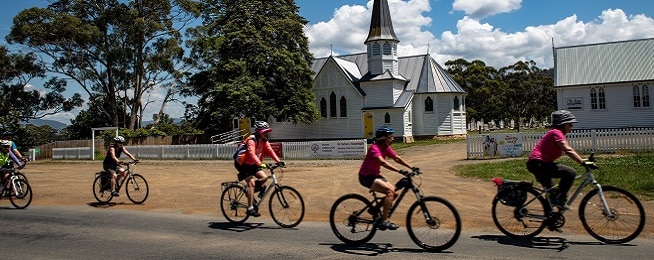 A group of people ride on a road past a white wooden church.