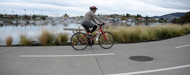 Woman riding a red bike along a concrete path with an overcast sky and yachts in Kangaroo Bay in the background.