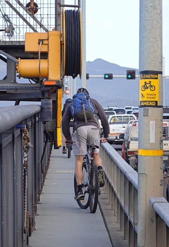 A man rides a bicycle over the Tasman Bridge - the photo shows the narrowness of the path, and a sign warning of a pinchpoint in front of him.