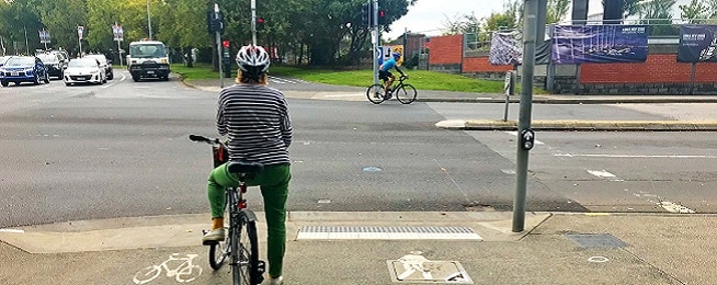 A woman wearing casual clothes waits at the lights on her bike while a road cyclist rides in front of her and traffic also waits at the lights.