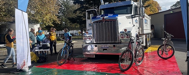 A large white track is surrounded by a colourful mat on the ground, with several bikes placed around the truck. A few people stand next to the truck talking.