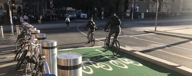 People ride in the separated cycleway on Swanston Street as it crosses Flinders Street in Melbourne, with green paint and white bicycle symbols in the foreground.