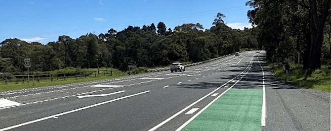 Image of road with green bicycle lane with painted buffer between it and the traffic lane, trees on either side of the road.